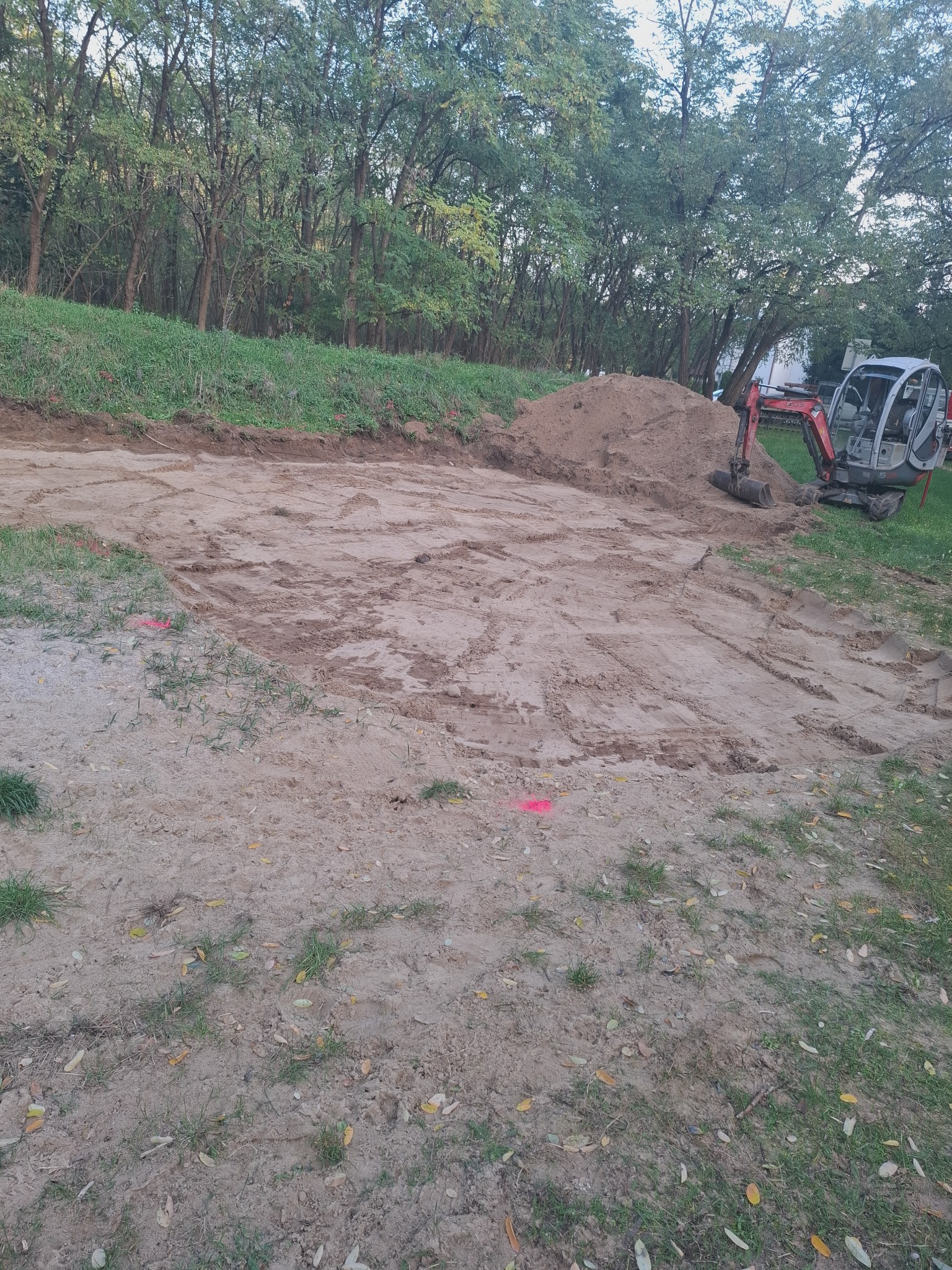 Teren wyrównany koparką, przygotowany pod budowę, z widocznym stosem ziemi i oznaczeniami geodezyjnymi.