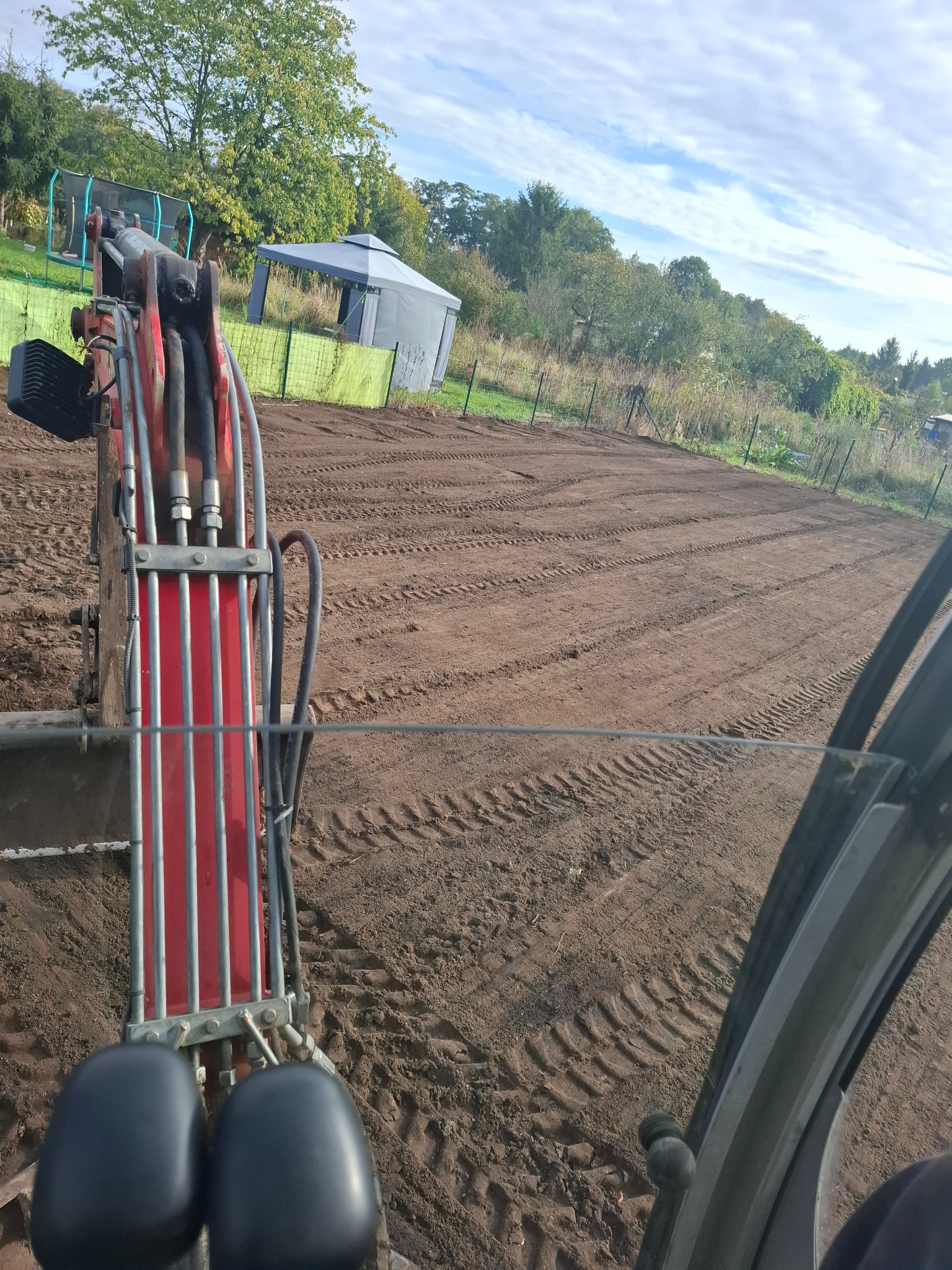 Widok z kabiny koparki na przygotowany teren pod uprawę, widoczne ślady opon i wyrównana ziemia w ogrodzie z trampoliną i altaną w tle.