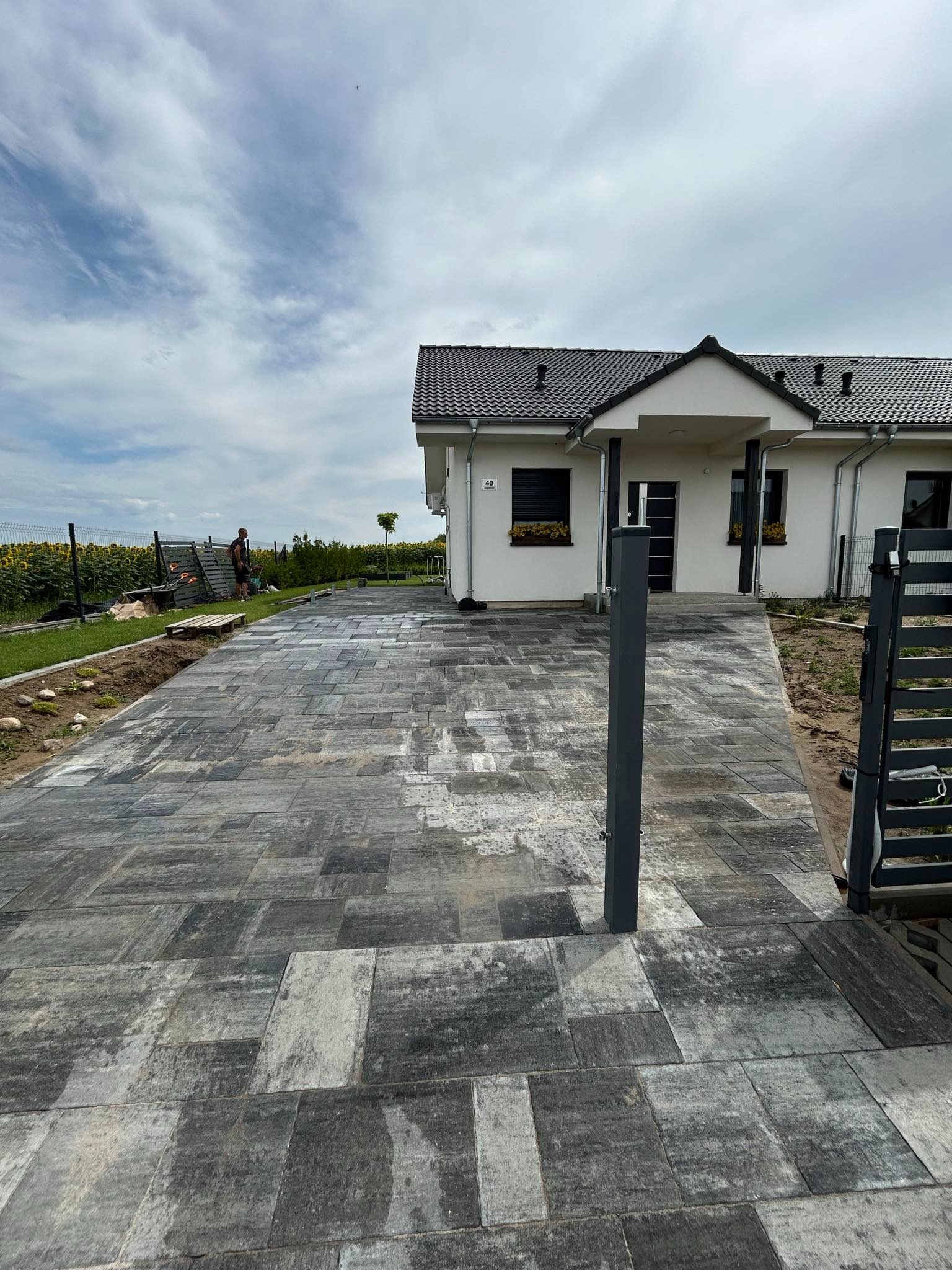 Ułożona kostka brukowa w odcieniach szarości prowadząca do nowoczesnego domu z ciemną dachówką, widoczny fragment metalowej bramy i osoba pracująca w tle przy ogrodzeniu.