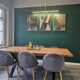 Jadalnia z drewnianym stołem na czarnych nogach, szare krzesła z pikowaniem i złotymi nogami, trzy obrazy w złotych ramach na ciemnozielonej ścianie, nowoczesna lampa sufitowa.