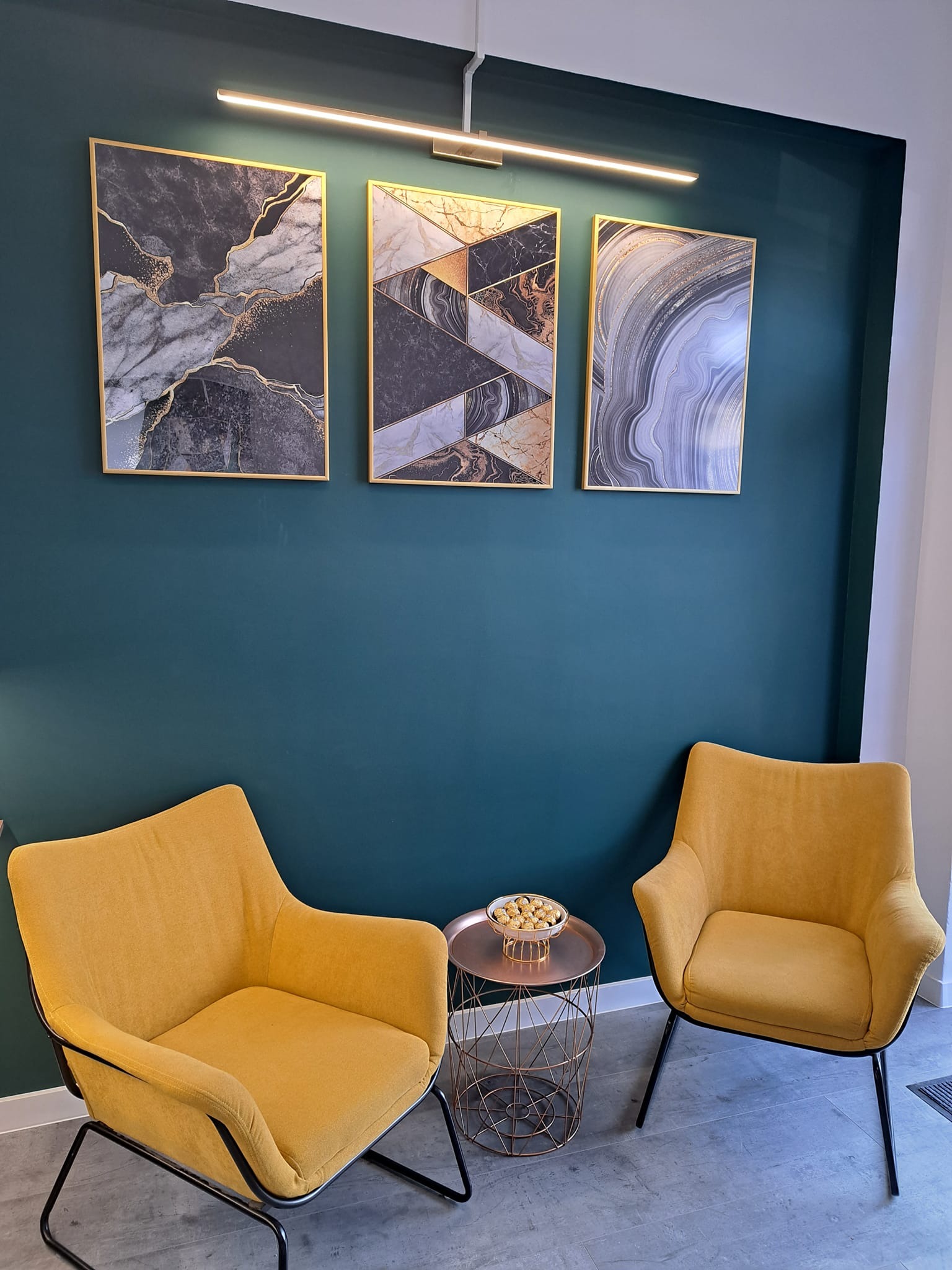 Pokój z dwoma żółtymi fotelami, okrągłym stolikiem z przekąskami i trzema abstrakcyjnymi obrazami na ciemnozielonej ścianie, oświetlony listwą LED.