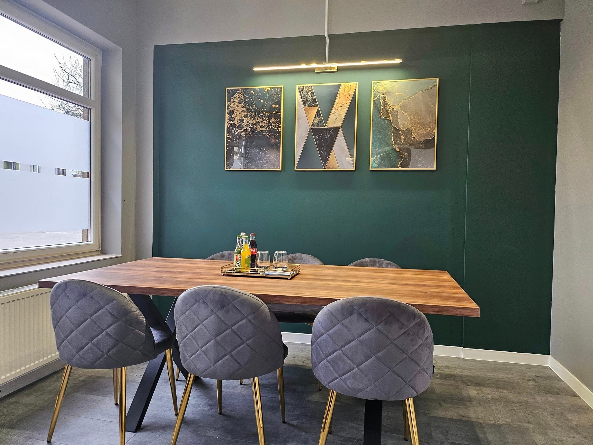 Jadalnia z drewnianym stołem na czarnych nogach, szare krzesła z pikowaniem i złotymi nogami, trzy obrazy w złotych ramach na ciemnozielonej ścianie, nowoczesna lampa sufitowa.
