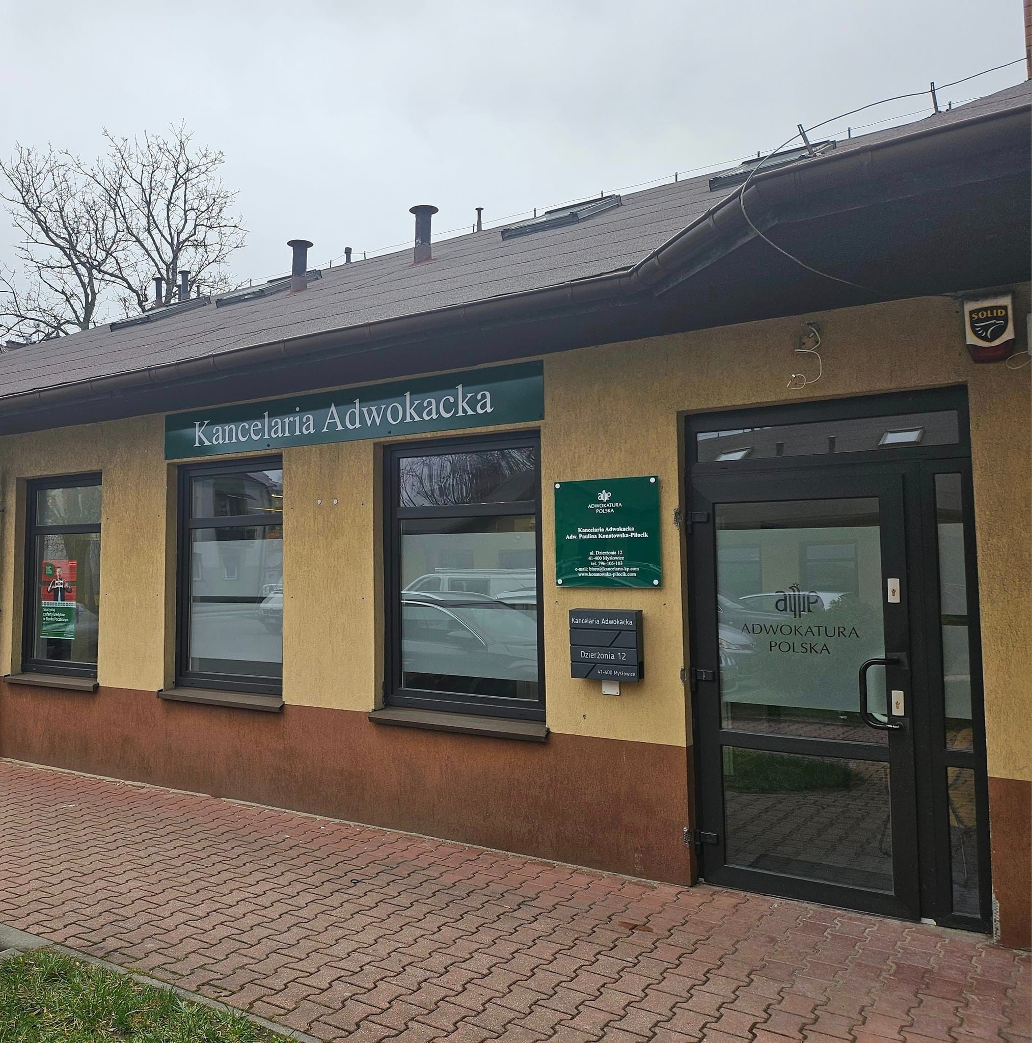 Zewnętrzny widok budynku Kancelarii Adwokackiej z szyldem, tabliczką informacyjną i skrzynką pocztową z adresem. Widoczne okna z plakatem oraz drzwi wejściowe z logo.