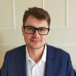 Portret prawnika w okularach i granatowym garniturze z poszetką w kropki na tle kremowej ściany z delikatną sztukaterią.