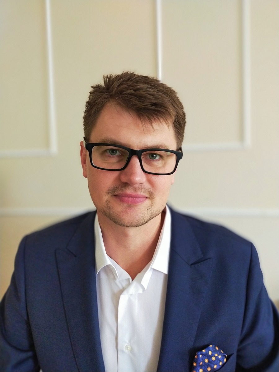 Portret prawnika w okularach i granatowym garniturze z poszetką w kropki na tle kremowej ściany z delikatną sztukaterią.