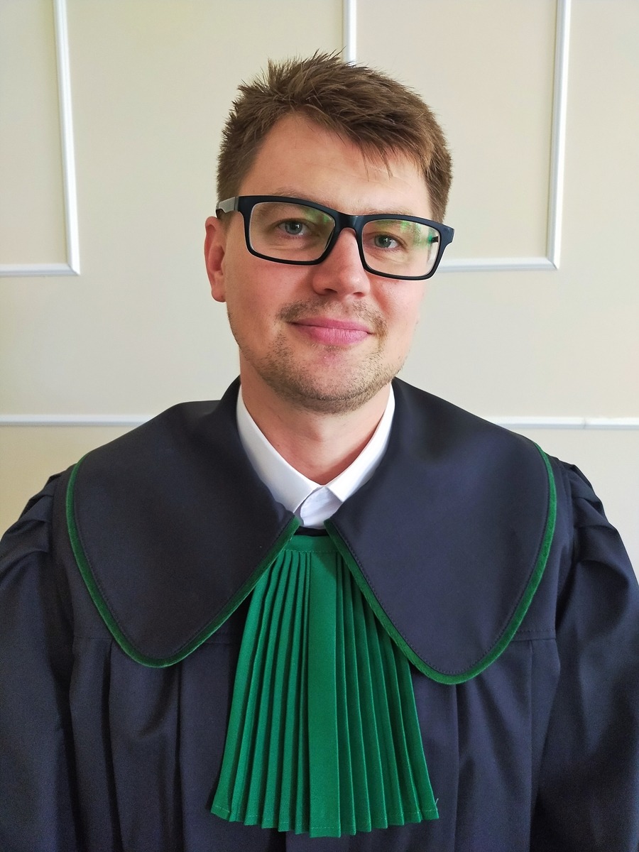 Portret adwokata w todze z zielonym żabotem, uśmiechnięty mężczyzna w okularach na tle jasnej ściany z dekoracyjną ramką.