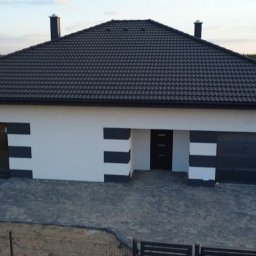 KamBud - Parterowy dom z ciemną dachówką i garażem, wykończony w biało-szare pasy. Ułożona kostka brukowa przed wejściem. Widok z góry, w tle drzewa.