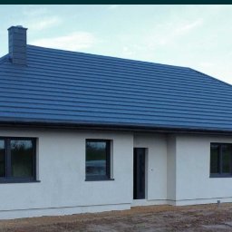 KamBud - Parterowy dom jednorodzinny z ciemną dachówką i garażem, widoczny z boku na tle zieleni i błękitnego nieba. Minimalistyczna bryła, nowoczesne okna.