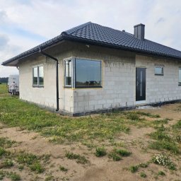 KamBud - Niedokończony dom z białych bloczków, czarny dach i okna z żółtą pianką montażową. Widoczny fragment lasu w tle i osoba przy białym samochodzie dostawczym.