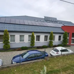 Szary budynek z panelami słonecznymi na dachu i czerwonym fragmentem elewacji. Przed budynkiem zaparkowane samochody i ozdobne drzewka.