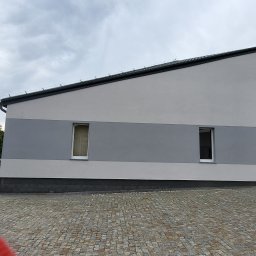 MatSolid-Bud - Dwukondygnacyjny budynek z szarą elewacją i dwoma oknami, widziany z poziomu brukowanego podjazdu w pochmurny dzień. Widoczny fragment zieleni i zaparkowany samochód.