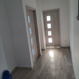 MatSolid-Bud - Wnętrze po remoncie: jasne ściany, drewnopodobne panele, dwie drewniane drzwi z przeszkleniami, fragment niebieskiego materiału w rogu.