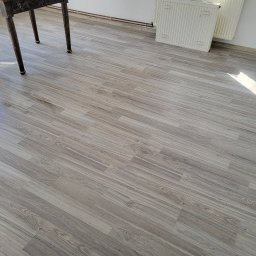 MatSolid-Bud - Ułożone panele podłogowe w odcieniach szarości, widoczne nogi stołu i fragment grzejnika. Ujęcie z góry, podkreślające wzór i strukturę paneli.