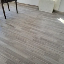 Ułożone panele podłogowe w odcieniach szarości, widoczne nogi stołu i fragment grzejnika. Ujęcie z góry, podkreślające wzór i strukturę paneli.