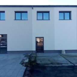 MatSolid-Bud - Jasna elewacja budynku z rzędem okien i drzwiami, widok z zewnątrz na szary chodnik. Minimalistyczny styl, równomierne oświetlenie.