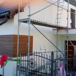 MatSolid-Bud - Dom w trakcie renowacji elewacji, widoczne rusztowania i fragment odnowionej ściany z drewnopodobną okładziną. Częściowo otynkowana ściana.