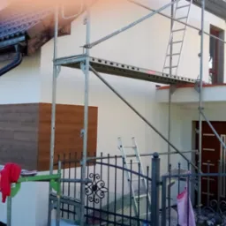 Dom w trakcie renowacji elewacji, widoczne rusztowania i fragment odnowionej ściany z drewnopodobną okładziną. Częściowo otynkowana ściana.