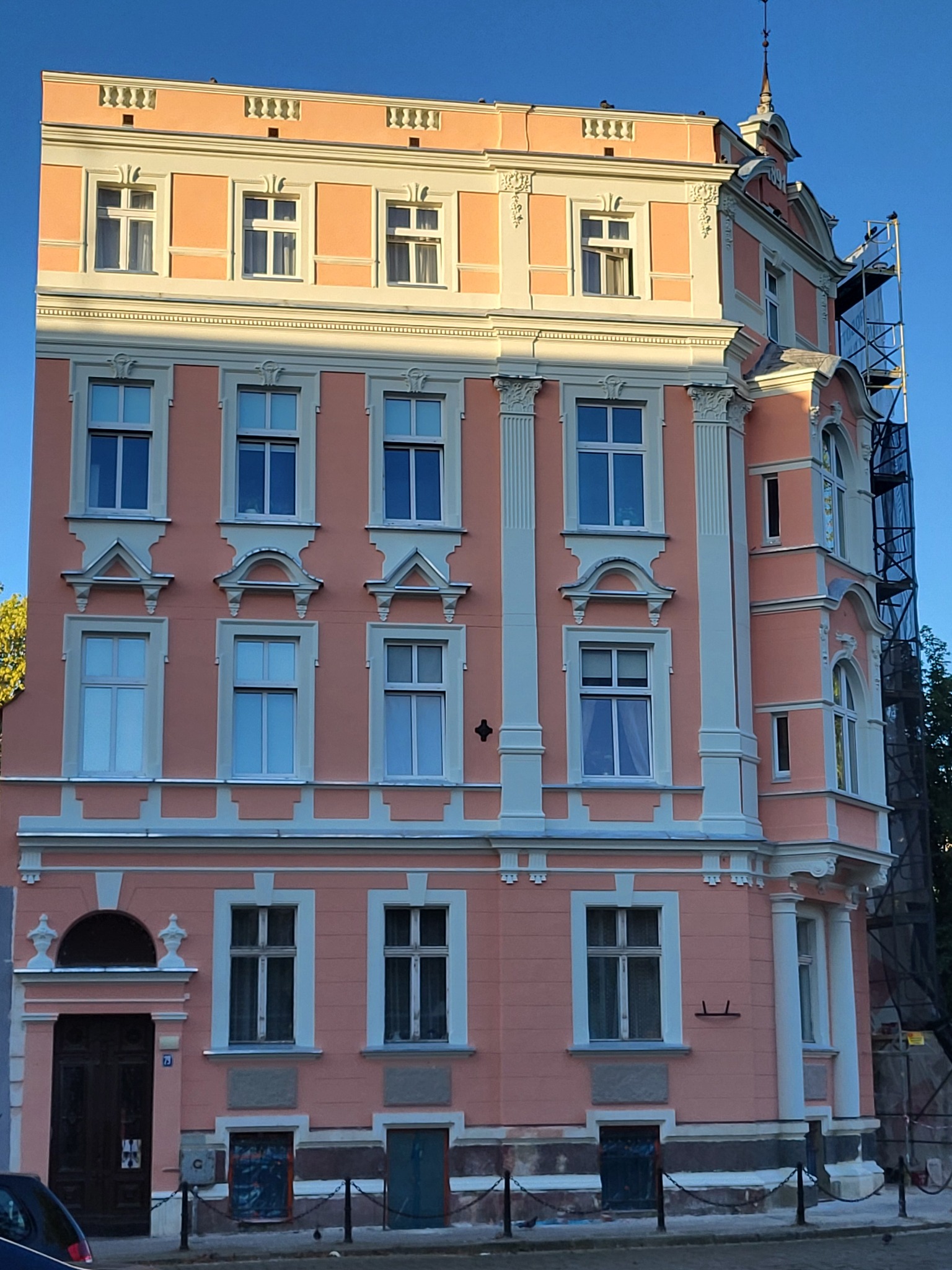 Odnowiona, różowa kamienica z białymi detalami architektonicznymi, widoczna z zewnątrz. W tle rusztowanie, sugerujące trwające prace konserwatorskie.