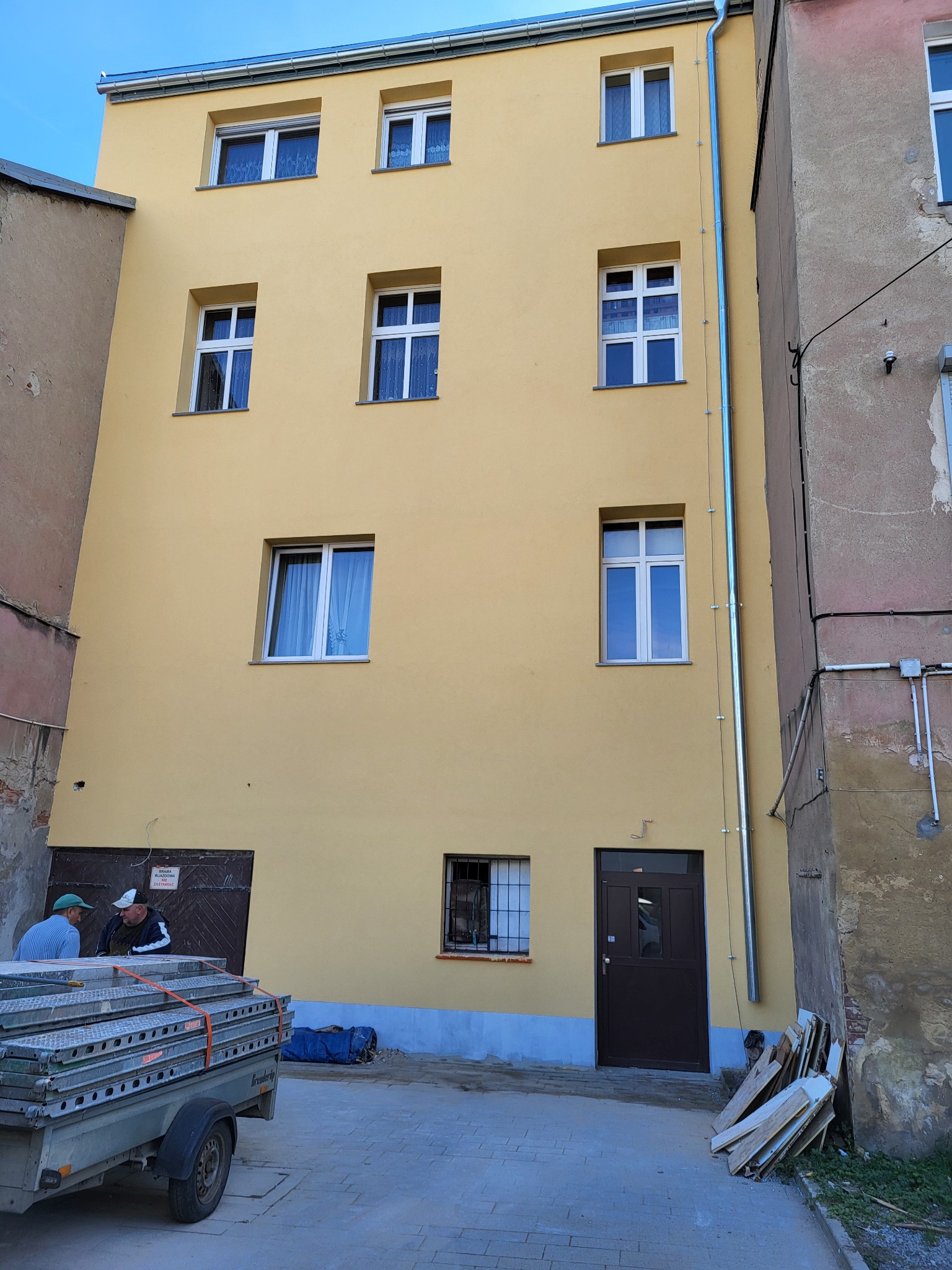 Żółta elewacja budynku z oknami i rynną, przy której stoją pracownicy i przyczepa ze sprzętem. Widoczne są elementy rusztowania i materiały budowlane.