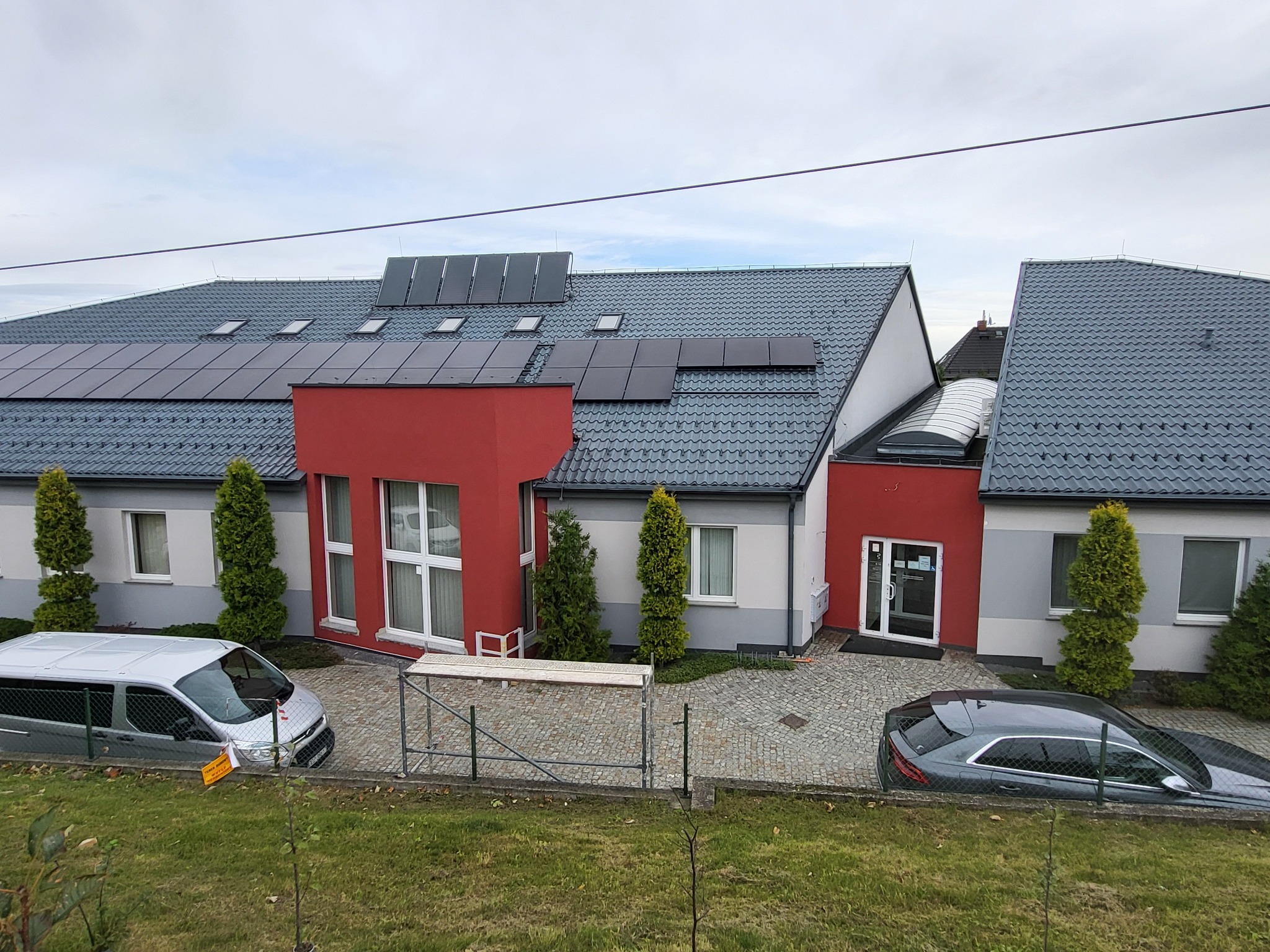 Budynek po renowacji elewacji z panelami słonecznymi na dachu, widoczny z góry. Na podwórku zaparkowane samochody i rozstawione rusztowanie.