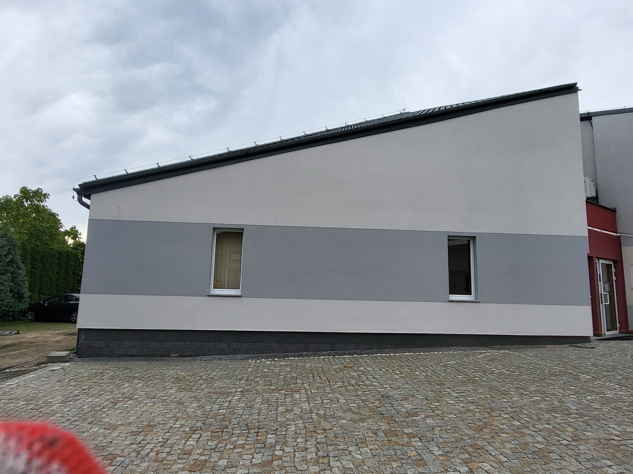 Dwukondygnacyjny budynek z szarą elewacją i dwoma oknami, widziany z poziomu brukowanego podjazdu w pochmurny dzień. Widoczny fragment zieleni i zaparkowany samochód.