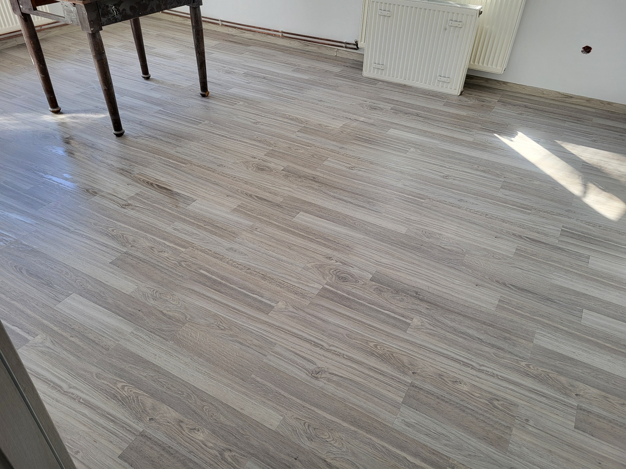 Ułożone panele podłogowe w odcieniach szarości, widoczne nogi stołu i fragment grzejnika. Ujęcie z góry, podkreślające wzór i strukturę paneli.