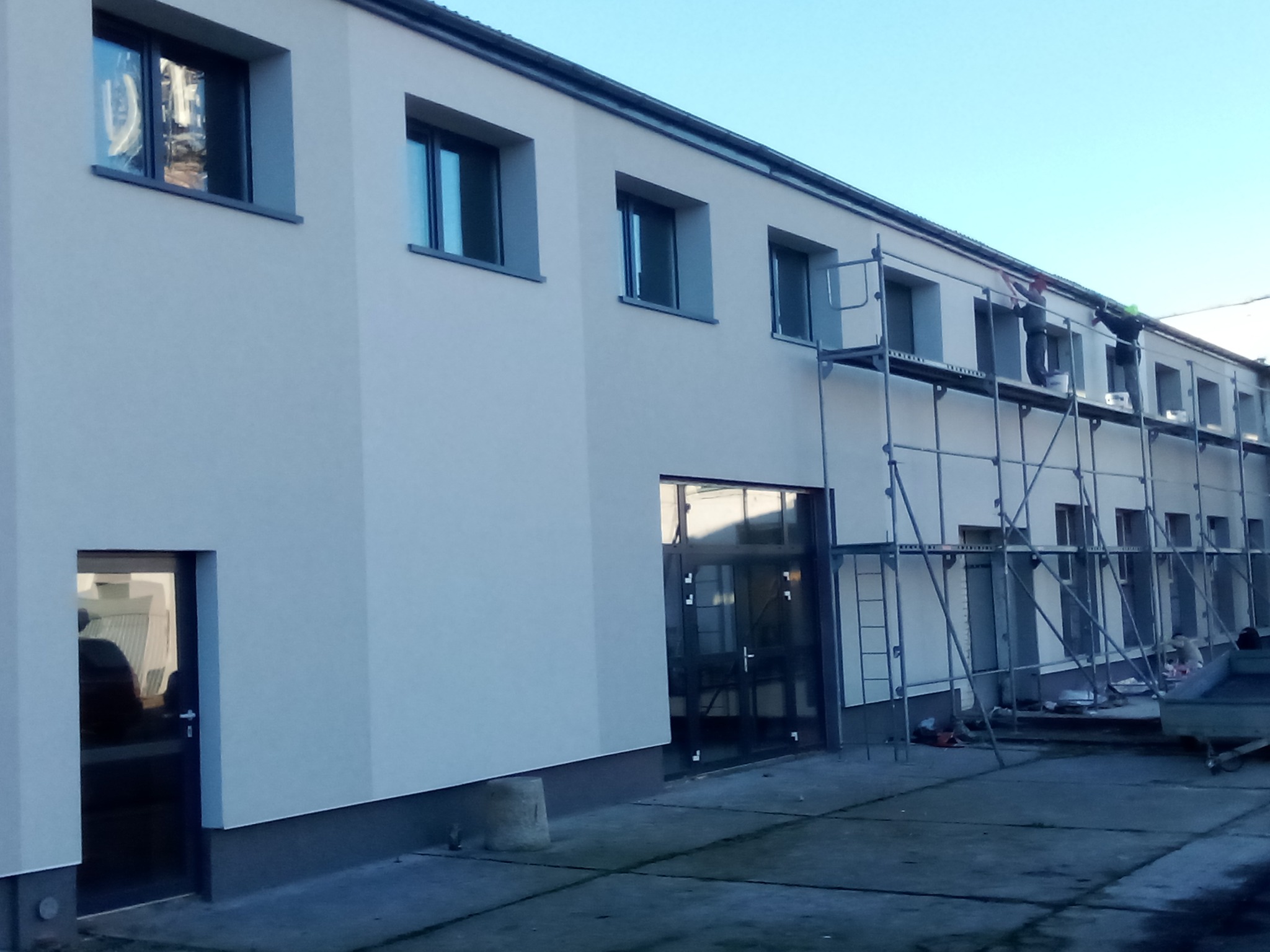 Budynek po renowacji elewacji, z rusztowaniami i pracownikami podczas malowania. Widoczne okna i drzwi, świeża, jasna elewacja.