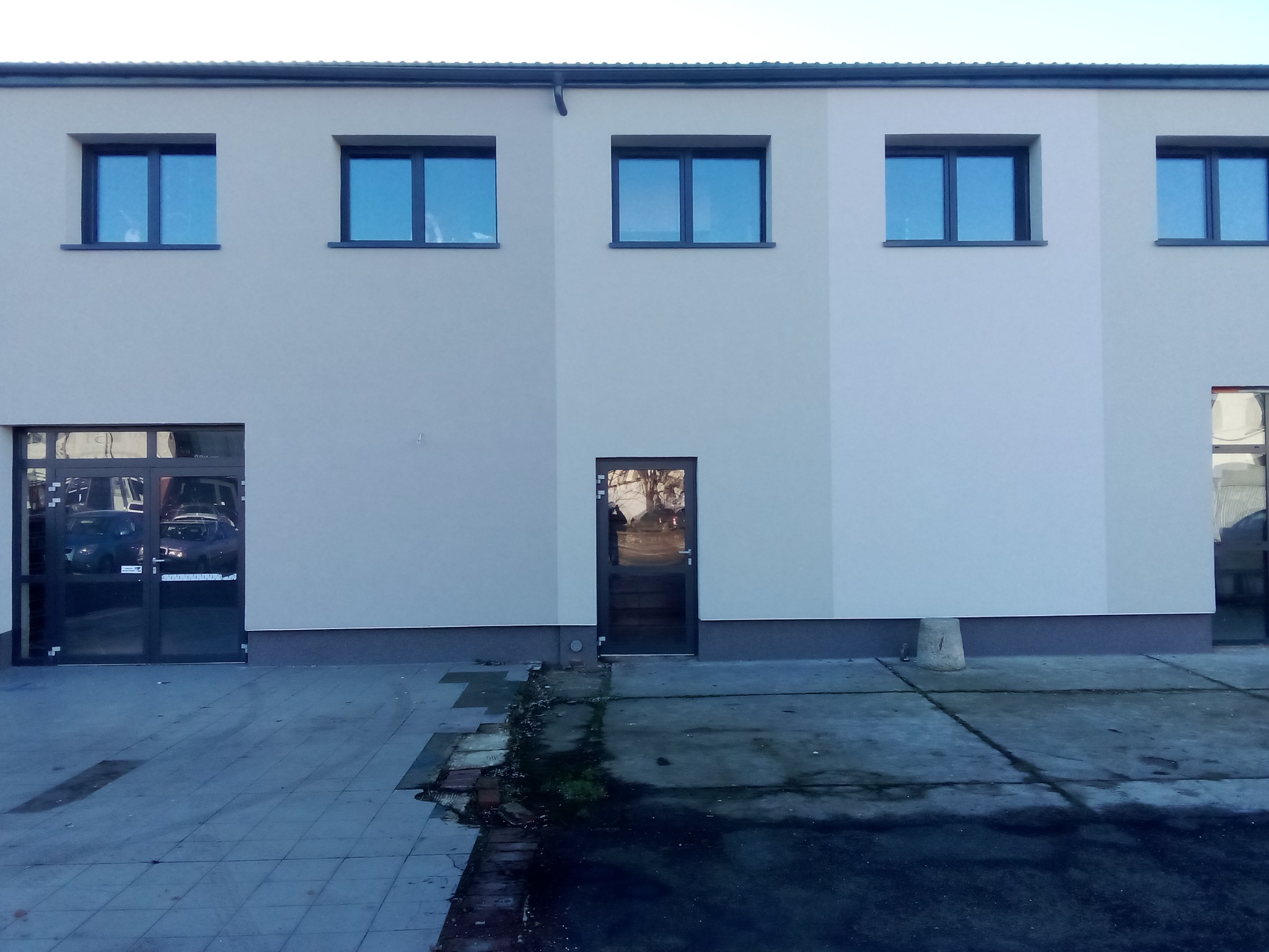 Jasna elewacja budynku z rzędem okien i drzwiami, widok z zewnątrz na szary chodnik. Minimalistyczny styl, równomierne oświetlenie.