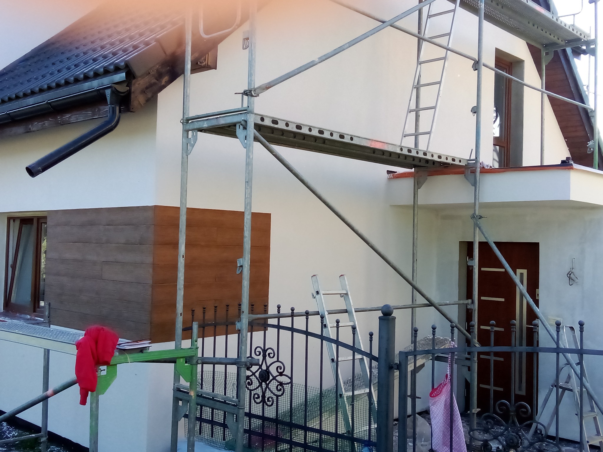 Dom w trakcie renowacji elewacji, widoczne rusztowania i fragment odnowionej ściany z drewnopodobną okładziną. Częściowo otynkowana ściana.