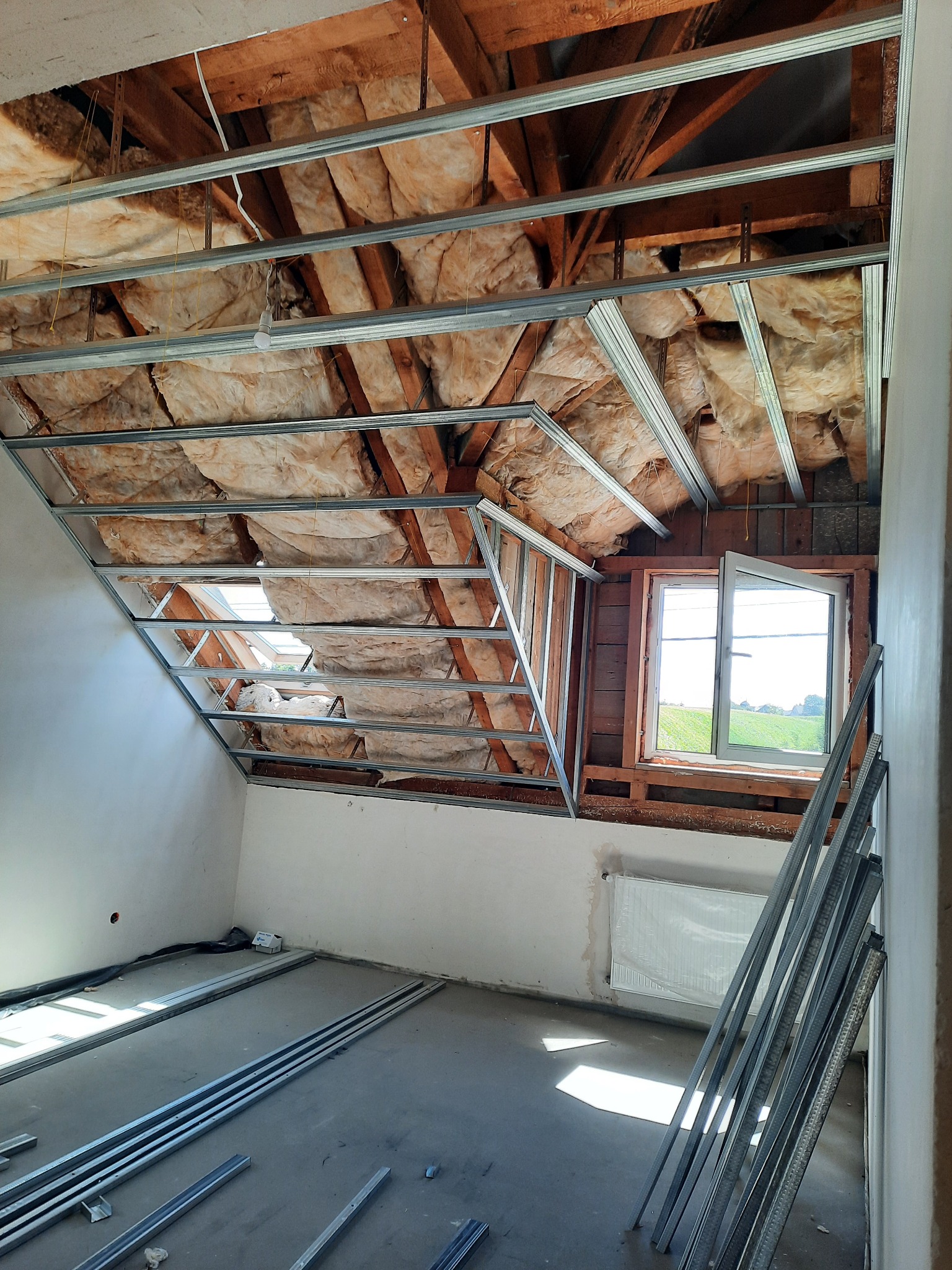 Poddasze ze skosami w trakcie ocieplania: widoczna wełna mineralna, stelaż metalowy pod płyty gipsowo-kartonowe i okno dachowe z widokiem na zewnątrz.