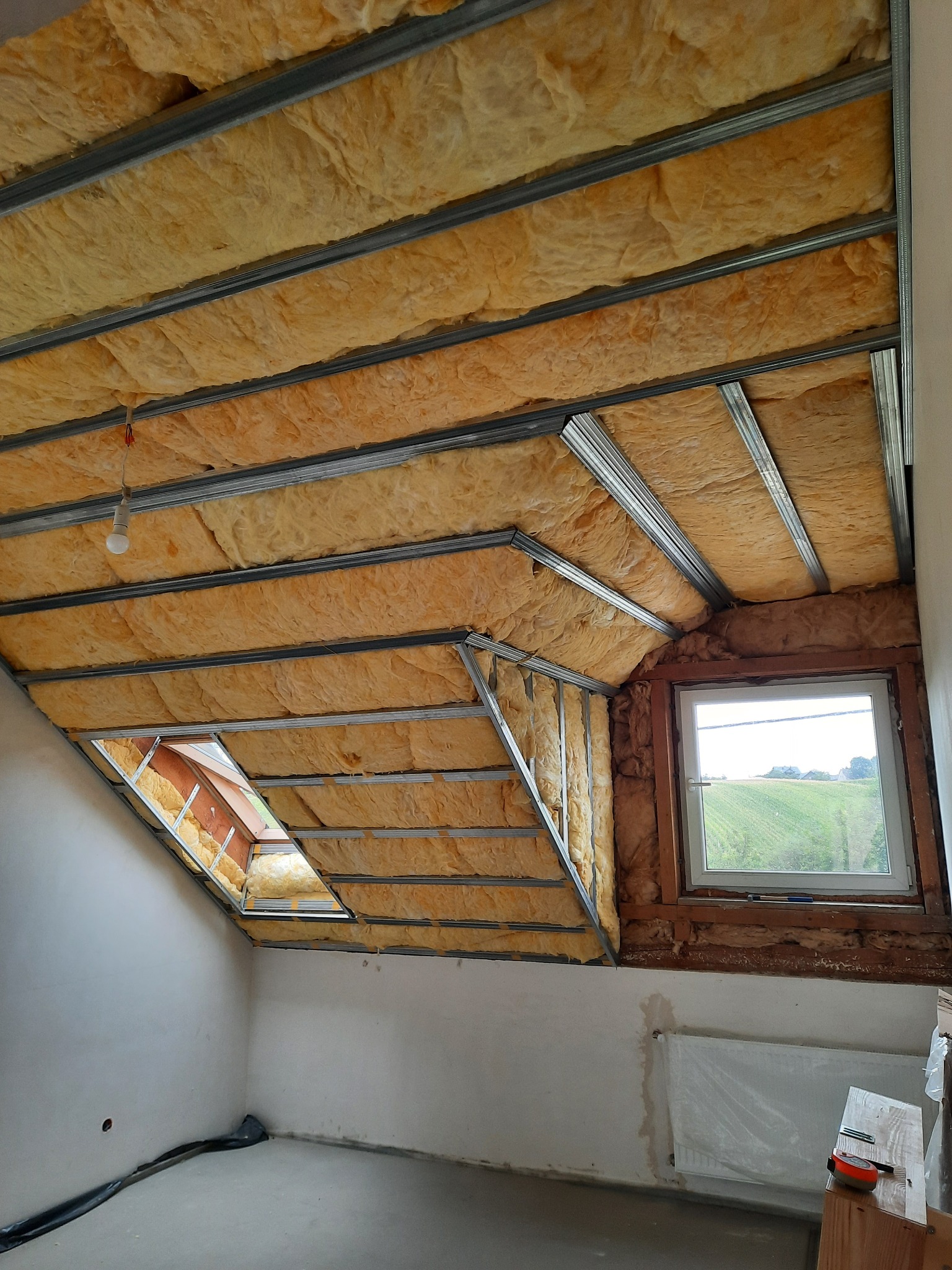 Poddasze w trakcie izolacji wełną mineralną, widoczne metalowe profile konstrukcyjne, okno dachowe i zwykłe okno z widokiem na zielone pole.