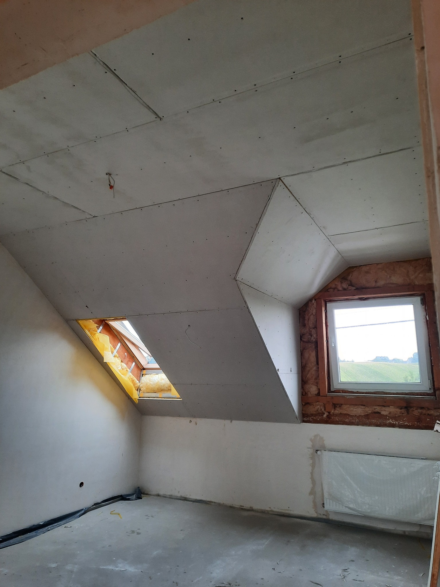 Poddasze w trakcie prac wykończeniowych: zamontowane płyty gipsowo-kartonowe na skosach dachu, widoczne okno dachowe z folią ochronną i fragment ocieplenia, okno pionowe z widokiem na zieleń...