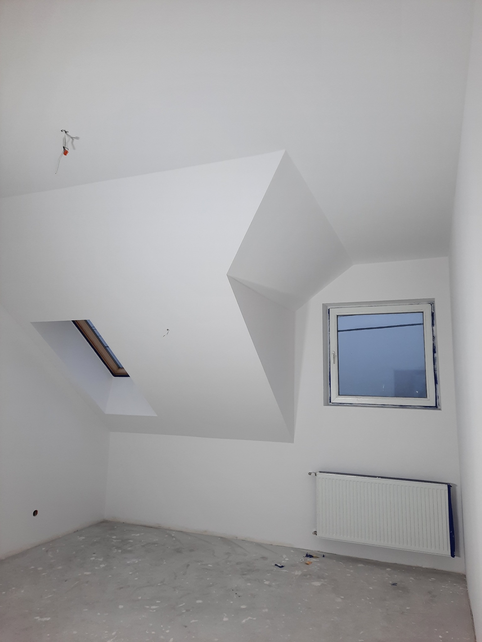 Poddasze po świeżym pomalowaniu na biało, widoczne okno dachowe i grzejnik, podłoga w trakcie przygotowania do dalszych prac wykończeniowych, surowy stan.
