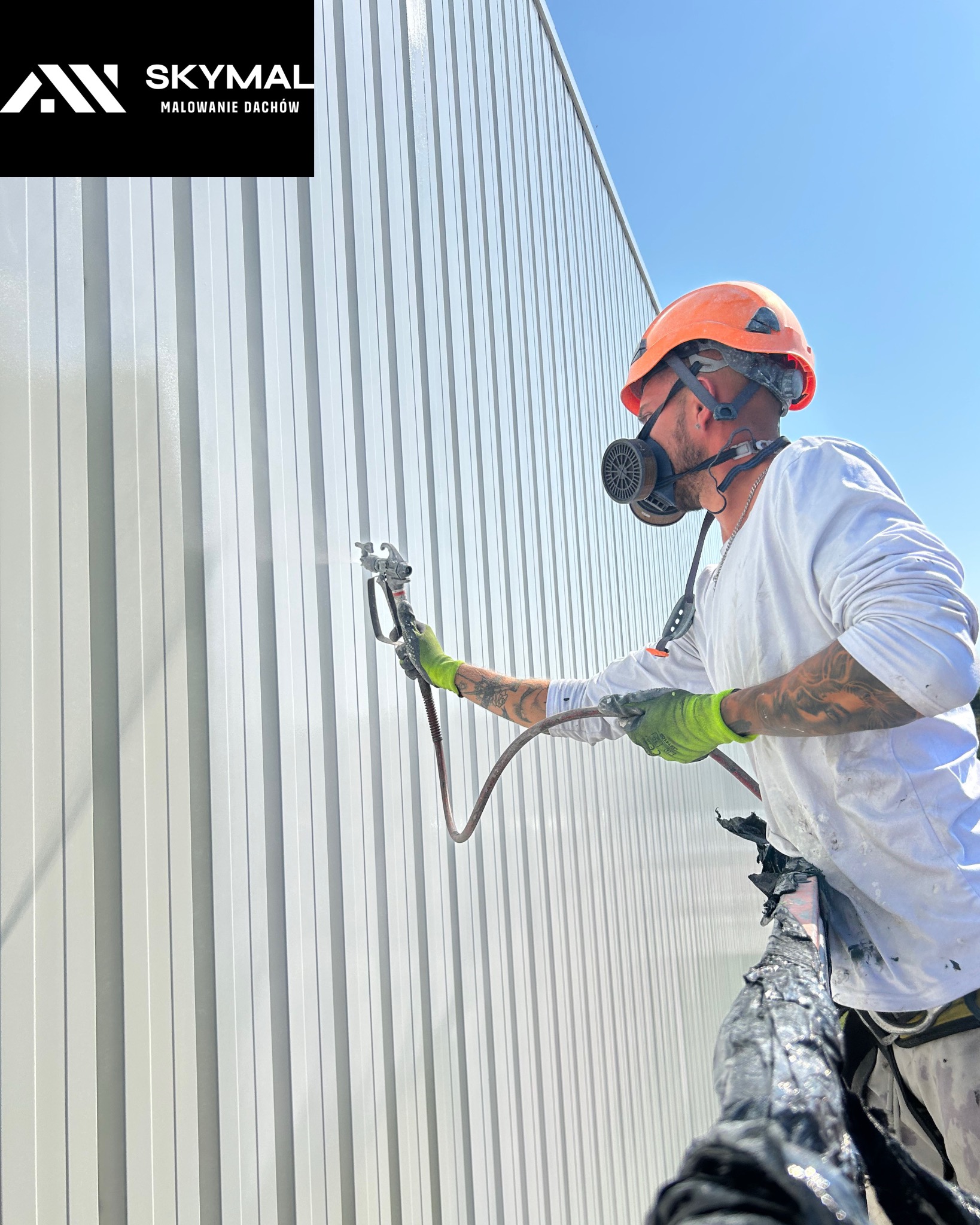 Pracownik w kasku i masce maluje natryskowo jasnoszarą, metalową elewację budynku. Widoczny pistolet natryskowy i świeżo pomalowana powierzchnia.