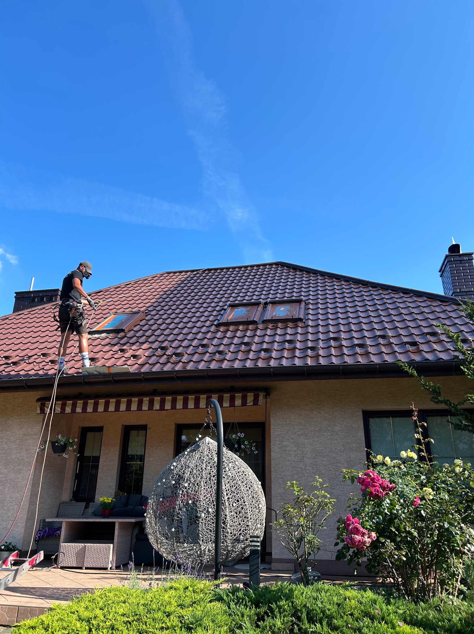 Pracownik w masce maluje okno dachowe na brązowym dachu domu jednorodzinnego. Widoczne narzędzia i lina zabezpieczająca. Błękitne niebo w tle.