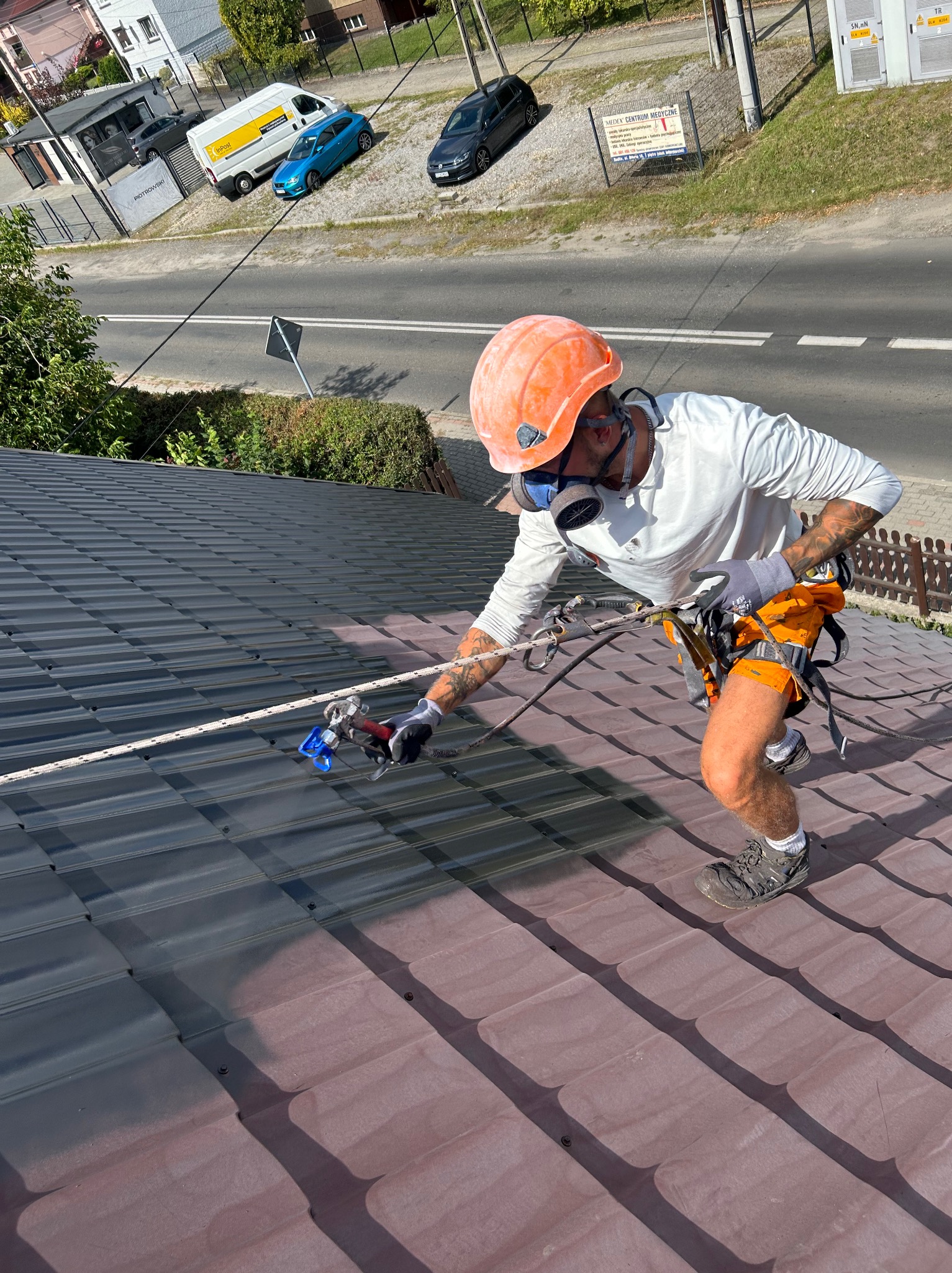 Pracownik w kasku i masce maluje dach metodą natryskową. Widoczna różnica koloru między pomalowaną a niepomalowaną częścią dachu. Praca na wysokości, zabezpieczenia linowe.