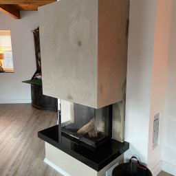 R S Budownictwo - Nowoczesny kominek z betonową obudową, czarnym blatem i paleniskiem z drewnem, widoczny kosz na drewno obok, na podłodze panele.