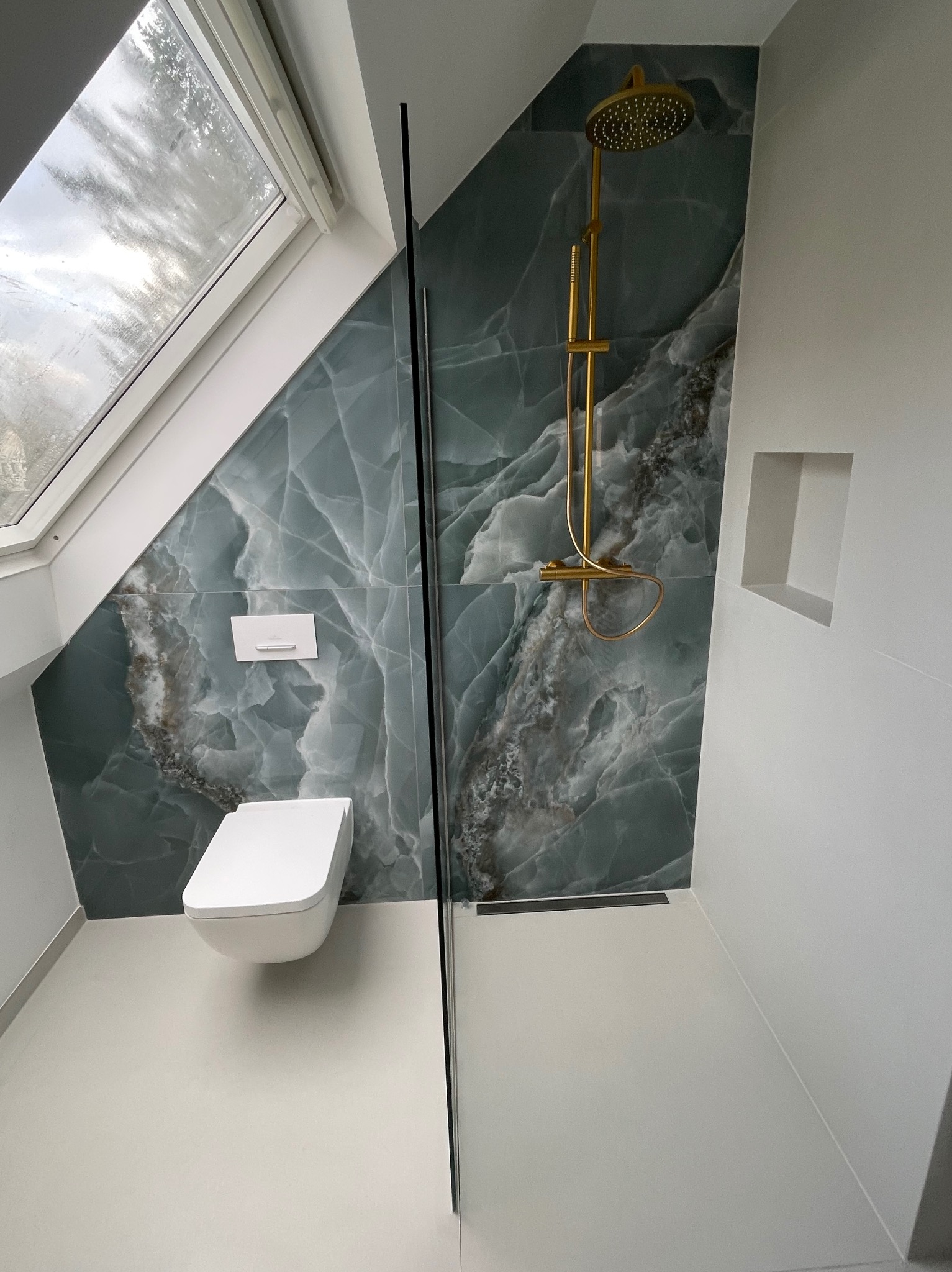 Nowoczesna łazienka na poddaszu z oknem dachowym, wykończona płytkami imitującymi marmur w odcieniach szarości i zieleni, z elementami armatury w kolorze złotym, kabiną prysznicową typu walk-in...