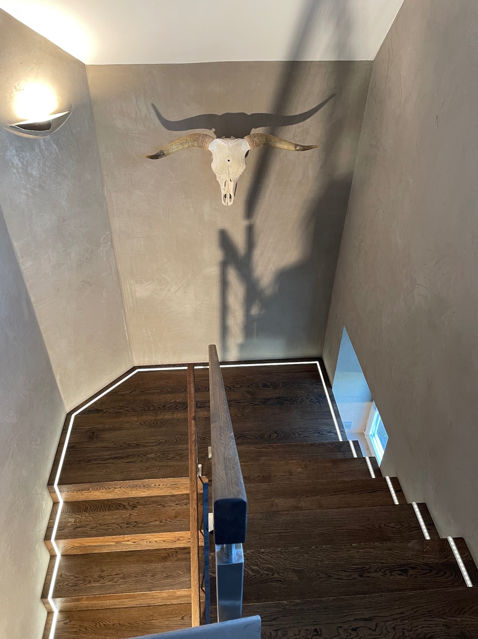 Klatka schodowa z drewnianymi stopniami oświetlonymi listwami LED, ściana wykończona w stylu betonu architektonicznego, dekoracja w postaci czaszki byka z rogami na ścianie.