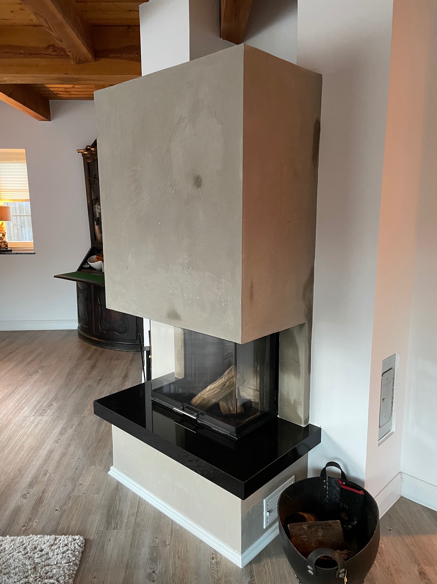 Nowoczesny kominek z betonową obudową, czarnym blatem i paleniskiem z drewnem, widoczny kosz na drewno obok, na podłodze panele.