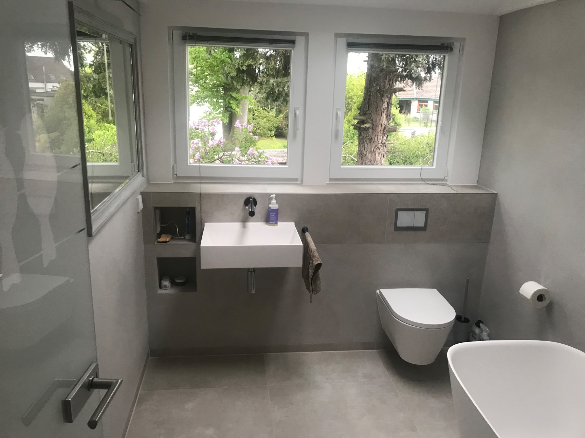 Nowoczesna łazienka w szarościach z wiszącą umywalką, toaletą i wanną, z widokiem na zieleń za oknem