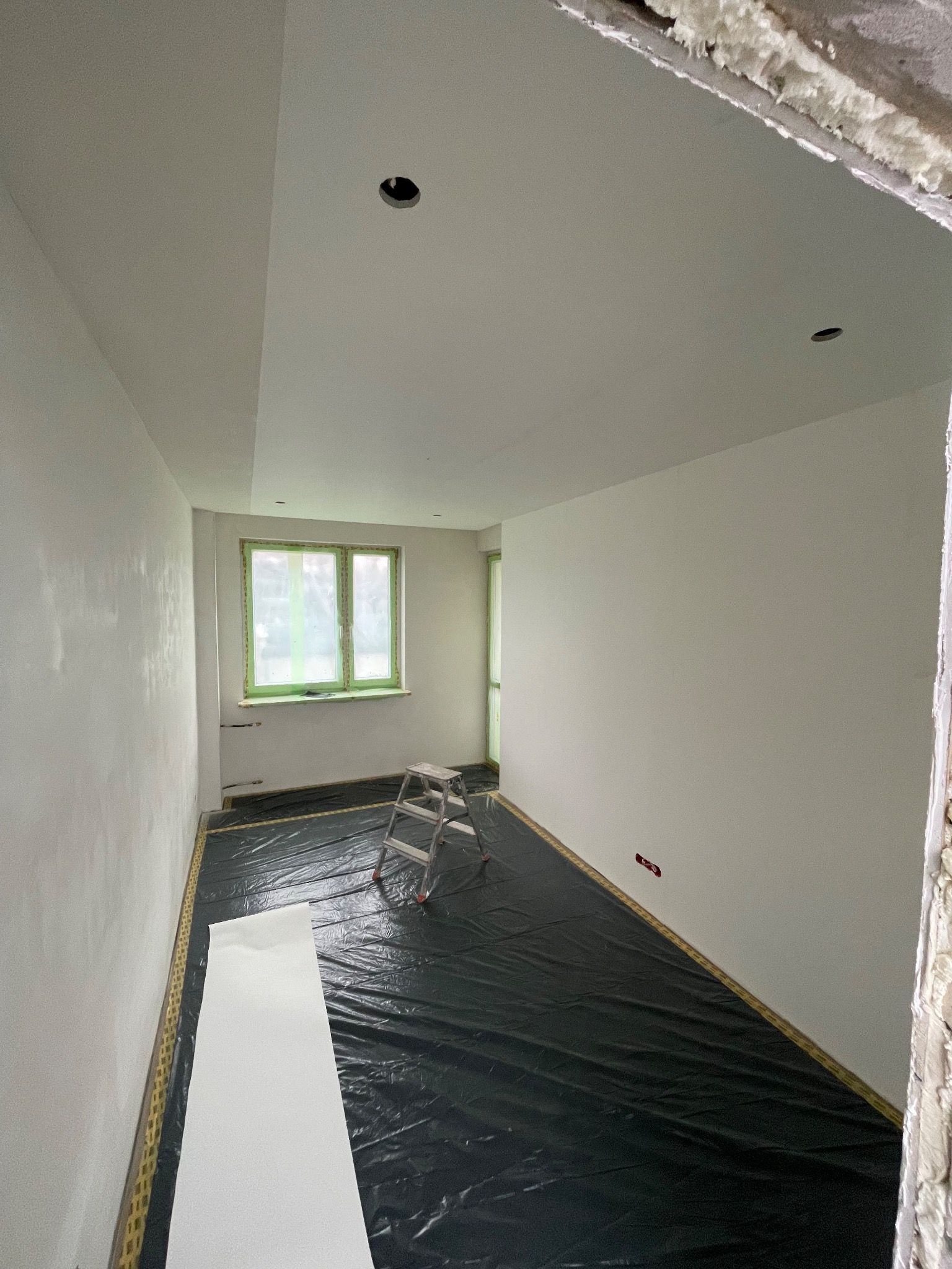 Przygotowane do malowania wnętrze pokoju w Łomży, z zabezpieczoną folią podłogą i oknem, widoczna drabina malarska i ślady gładzi na ścianach.