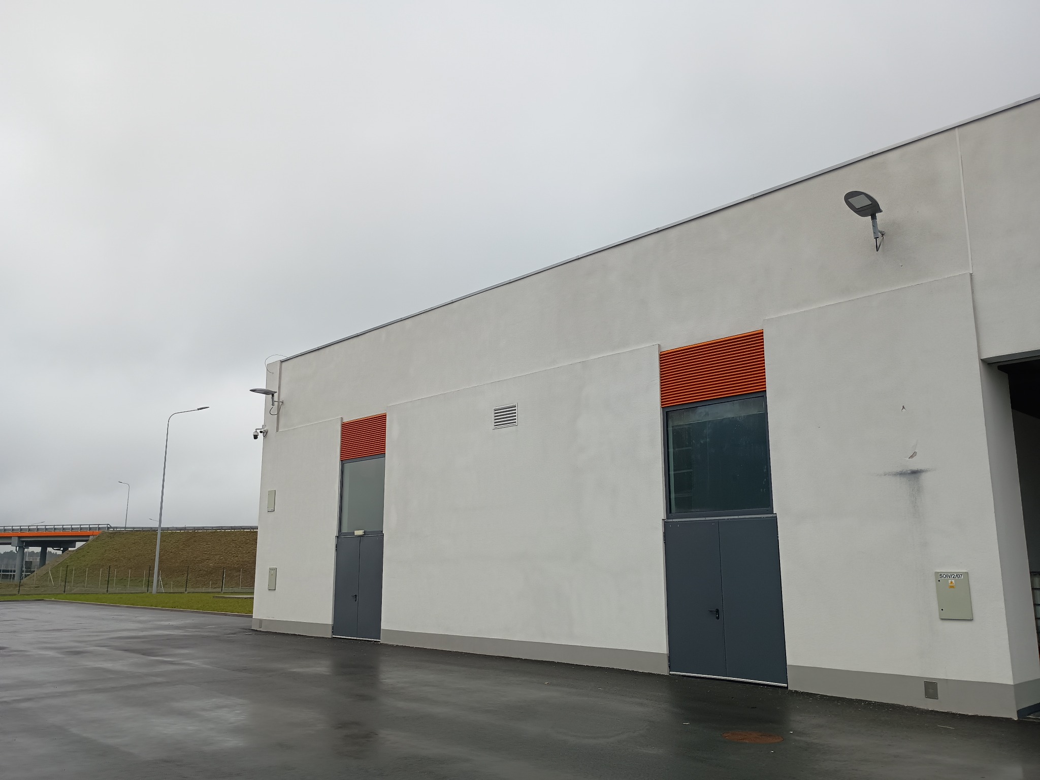 Elewacja budynku przemysłowego z dwoma parami drzwi i okien z pomarańczowymi żaluzjami, oświetleniem zewnętrznym LED i skrzynką elektryczną na ścianie, na tle pochmurnego nieba i wiaduktu.