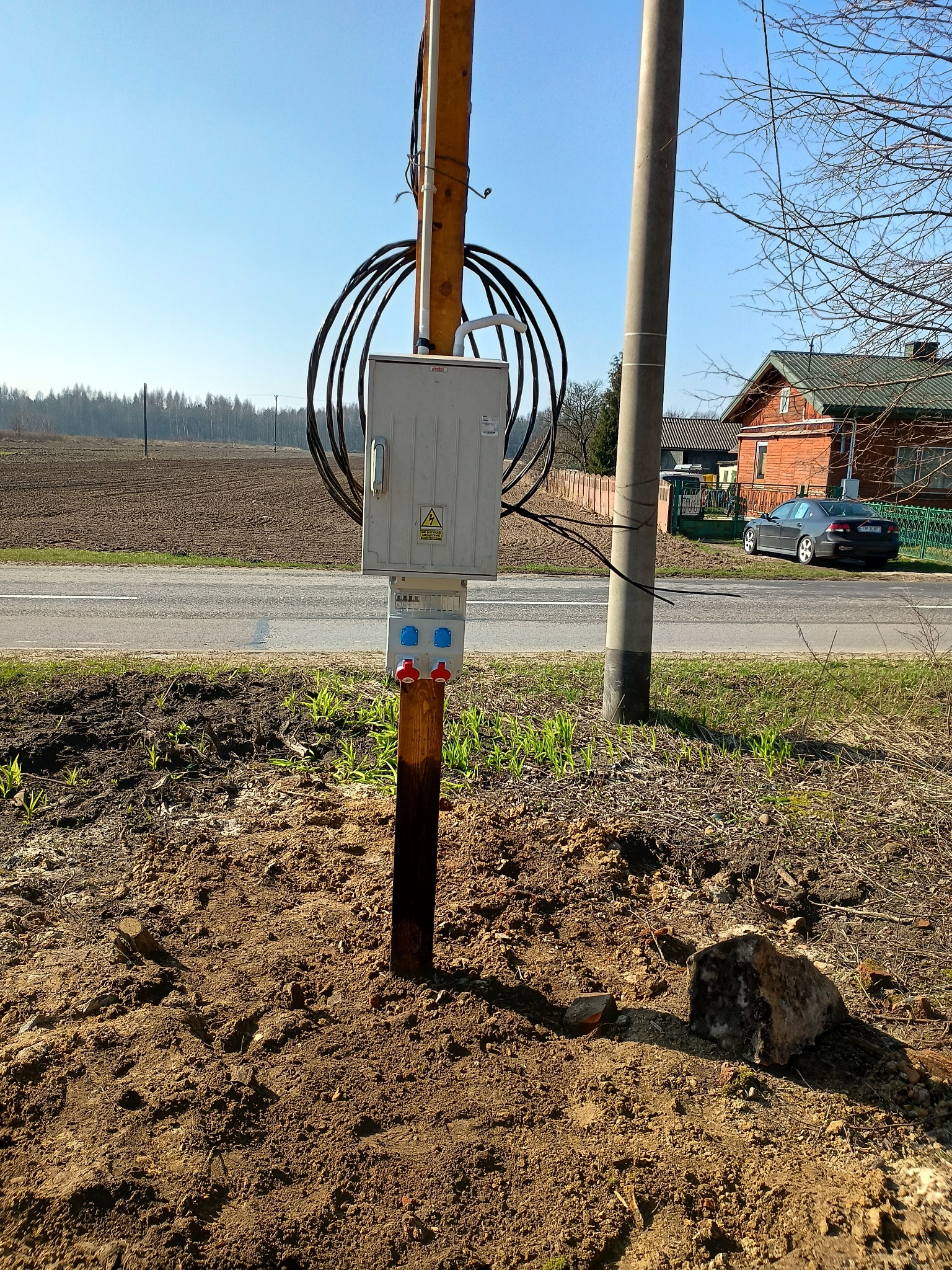 Słup z zamontowaną skrzynką elektryczną i zwisającymi kablami, umieszczony na poboczu drogi w wiejskim krajobrazie, widoczne gniazda z czerwonymi i niebieskimi wtyczkami, w tle pole i dom.