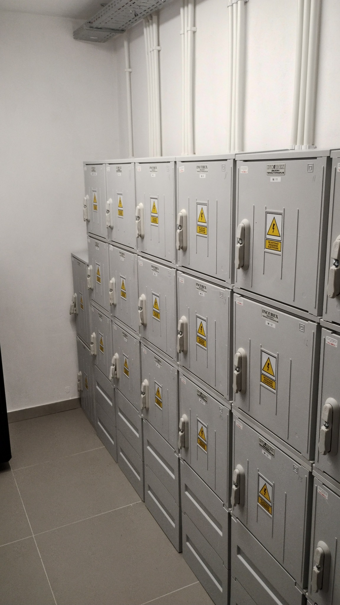 Rząd szarych skrzynek elektrycznych INCOBEX z ostrzeżeniami o napięciu, umieszczonych w rzędach wzdłuż ściany w pomieszczeniu technicznym, z widocznymi rurami instalacyjnymi pod sufitem.