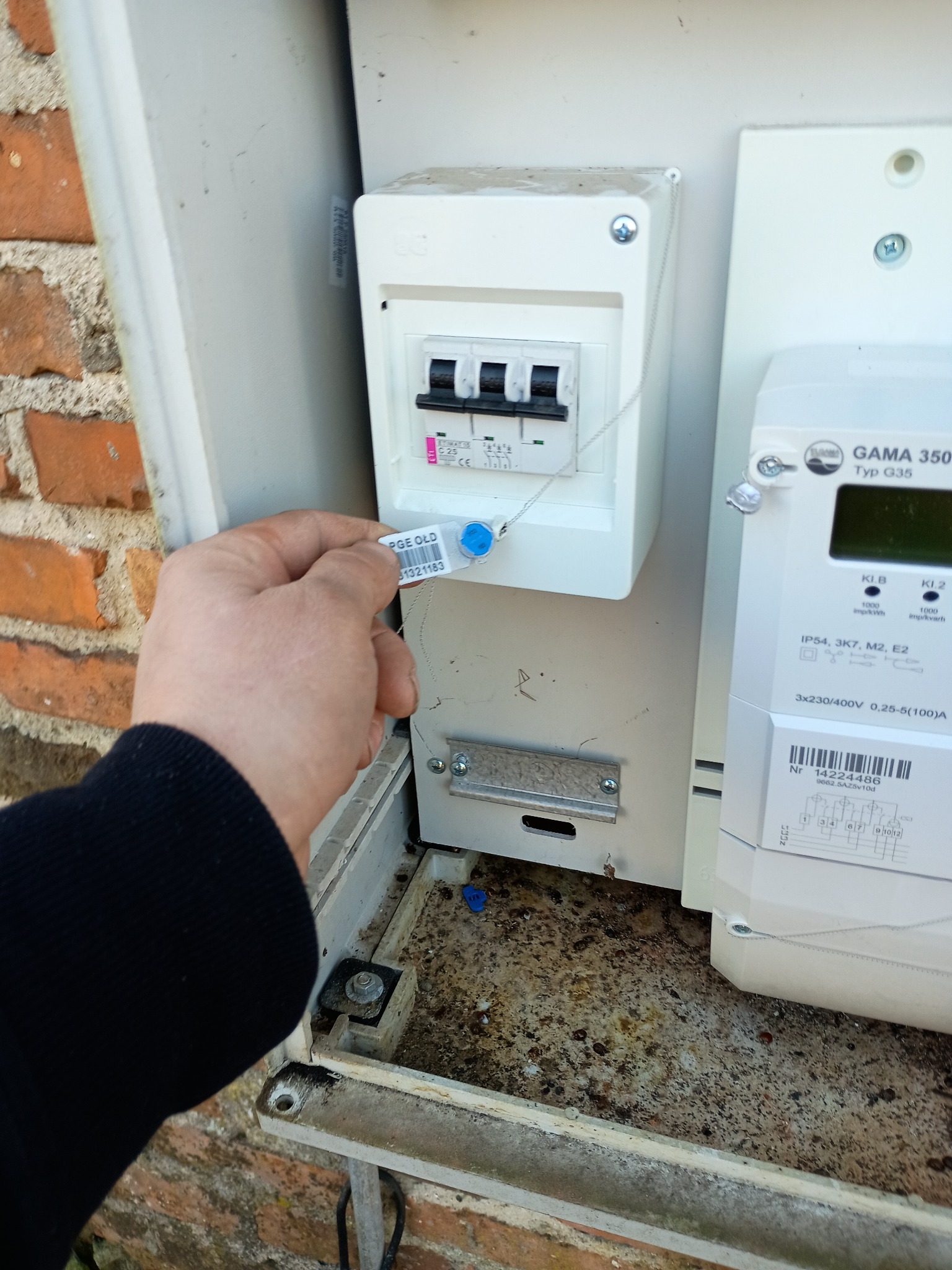 Zapinanie plomby na zabezpieczeniu w skrzynce elektrycznej, widoczny licznik energii elektrycznej GAMA 350 i fragment ceglanej ściany.