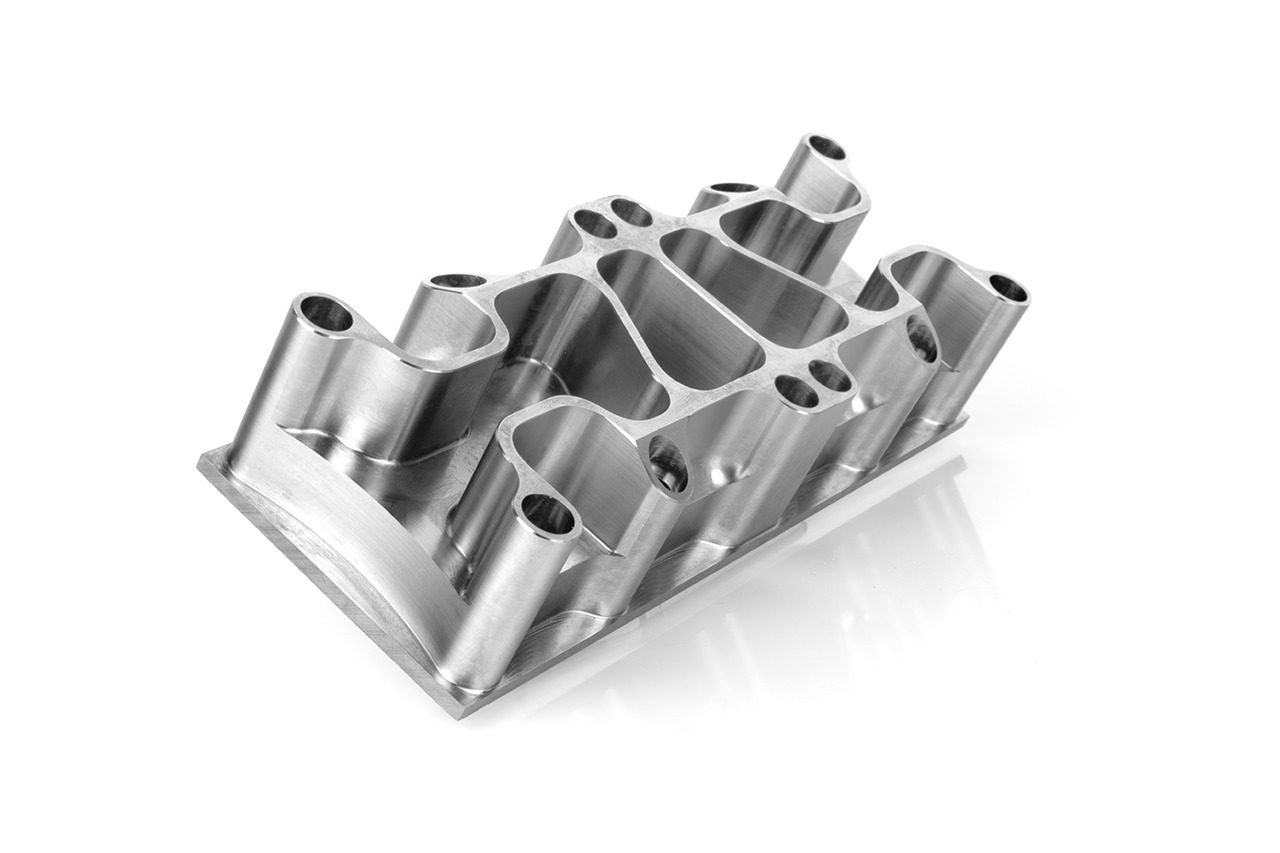 Precyzyjnie wykonany, aluminiowy element o skomplikowanej, ażurowej strukturze, z licznymi otworami i gładką, satynową powierzchnią, leżący na białym tle z widocznym odbiciem.