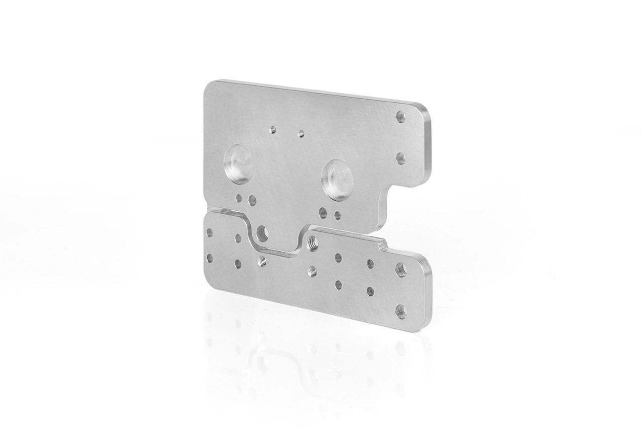 Precyzyjnie obrobiony, srebrny element metalowy o nieregularnym kształcie z licznymi otworami, widoczne gwintowane otwory, leżący na białym tle z odbiciem.