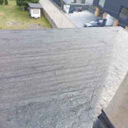 PATRYK FLORSZ - Płaski dach pokryty szarą, bitumiczną masą uszczelniającą, widok z góry na okoliczne budynki i ulicę.