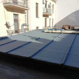 Płaski dach pokryty papą termozgrzewalną na budynku wielorodzinnym, widoczne detale łączeń i fragmenty konstrukcji dachu, w tle budynek z balkonami i instalacjami.